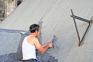  Ein Dachdecker schlägt mit seinem traditio­nellen Werkzeug – einem Hammer und einer Haubrücke –&nbsp;einen Schieferstein zu 